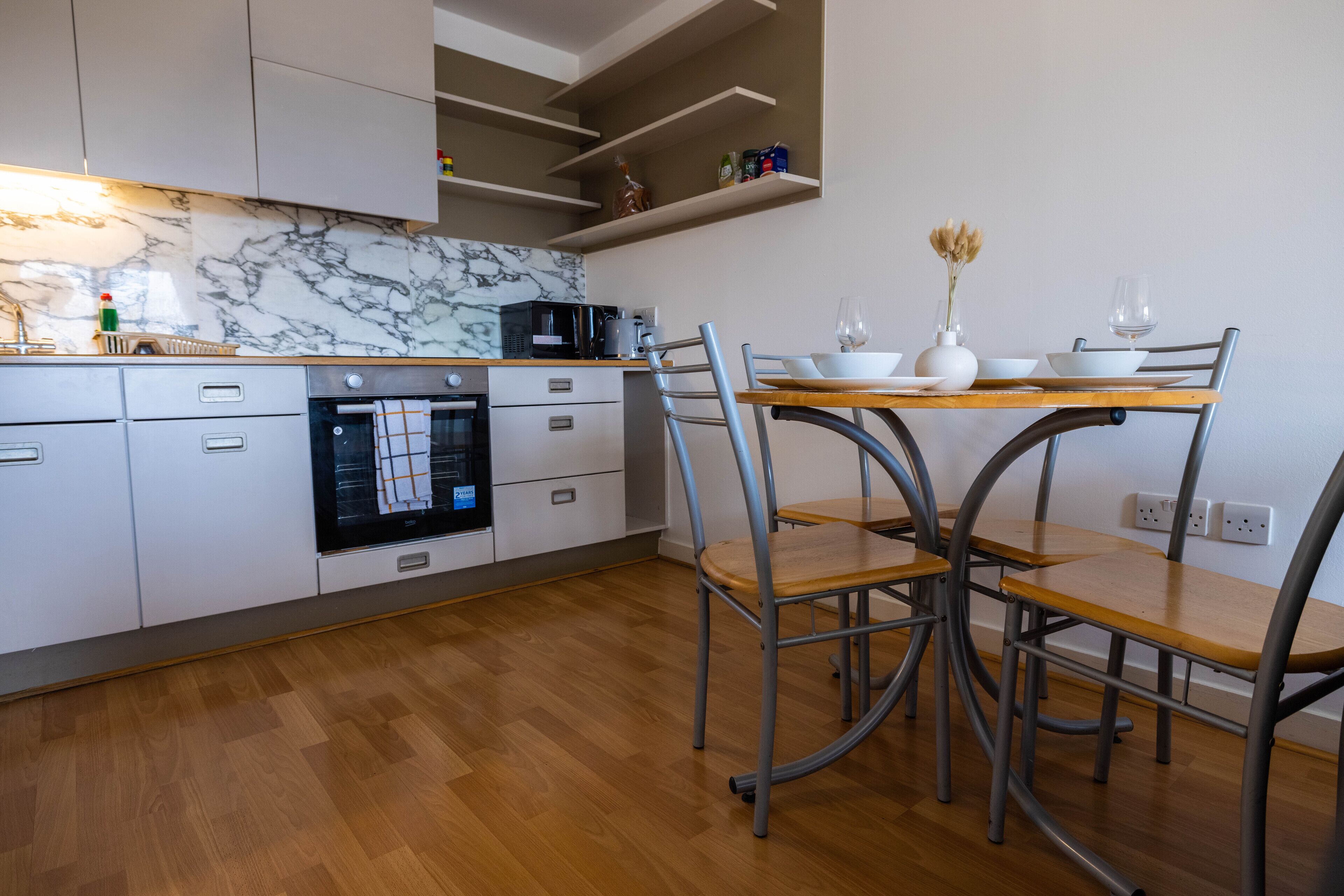 Une cuisine moderne avec des armoires blanches, un pare-feu en marbre, un four encastré et un coin repas comprenant une table en bois et des chaises en métal.