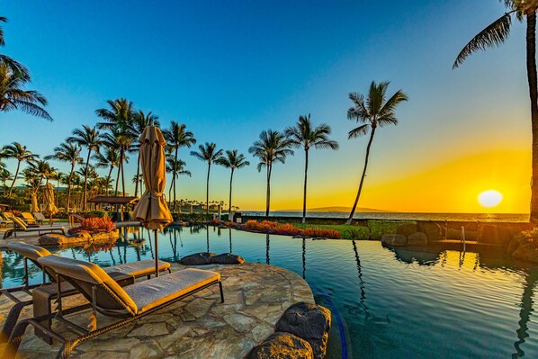 A resort pool area with lounge chairs and umbrellas, surrounded by palm trees and a sunset view.