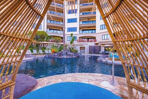 Zona de piscina con vista a un edificio de varios pisos, sillas de descanso y una cascada.