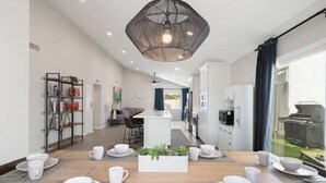 Une cuisine moderne dotée d’un îlot central, un coin repas avec de la vaisselle blanche, et un espace de vie comprenant une bibliothèque et un téléviseur.