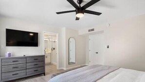 Une chambre à coucher comprenant un lit, une commode, un téléviseur, un ventilateur de plafond et une porte donnant sur une salle de bain.
