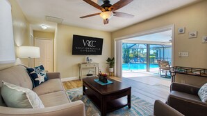 A living room with a ceiling fan, a TV displaying "VACATION RENTALS CAPE CORAL", and a view of a pool through glass doors. - VRCC Villa Paradise Palms (Cape Coral)