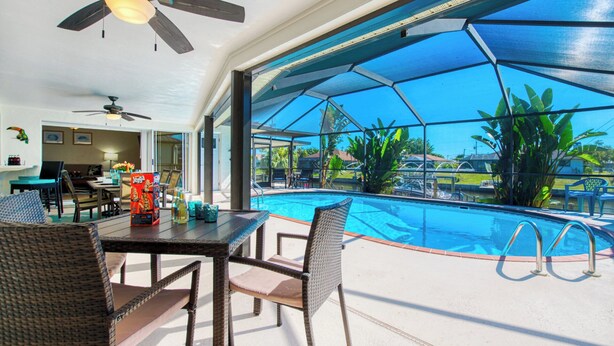 A poolside area with a glass-enclosed pool, wicker furniture, ceiling fans, and a view of the surrounding landscape. - VRCC Villa Paradise Palms (Cape Coral)