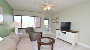 A living room with a beige sofa, a glass coffee table, a leather chair, a television on a white cabinet, and a ceiling fan. - Beautiful marina view in the Keel Club @ Burnt Store Marina and Country club (Punta Gorda)