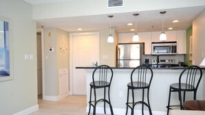A modern kitchen with a stainless steel refrigerator, a microwave, and a built-in oven. There are three black bar stools and pendant lights above the counter. - Beautiful marina view in the Keel Club @ Burnt Store Marina and Country club (Punta Gorda)