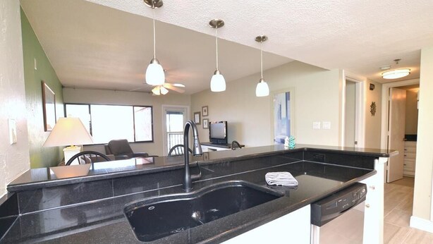 A kitchen with a black countertop, a sink, and a dishwasher. There are pendant lights above the counter. - Beautiful marina view in the Keel Club @ Burnt Store Marina and Country club (Punta Gorda)