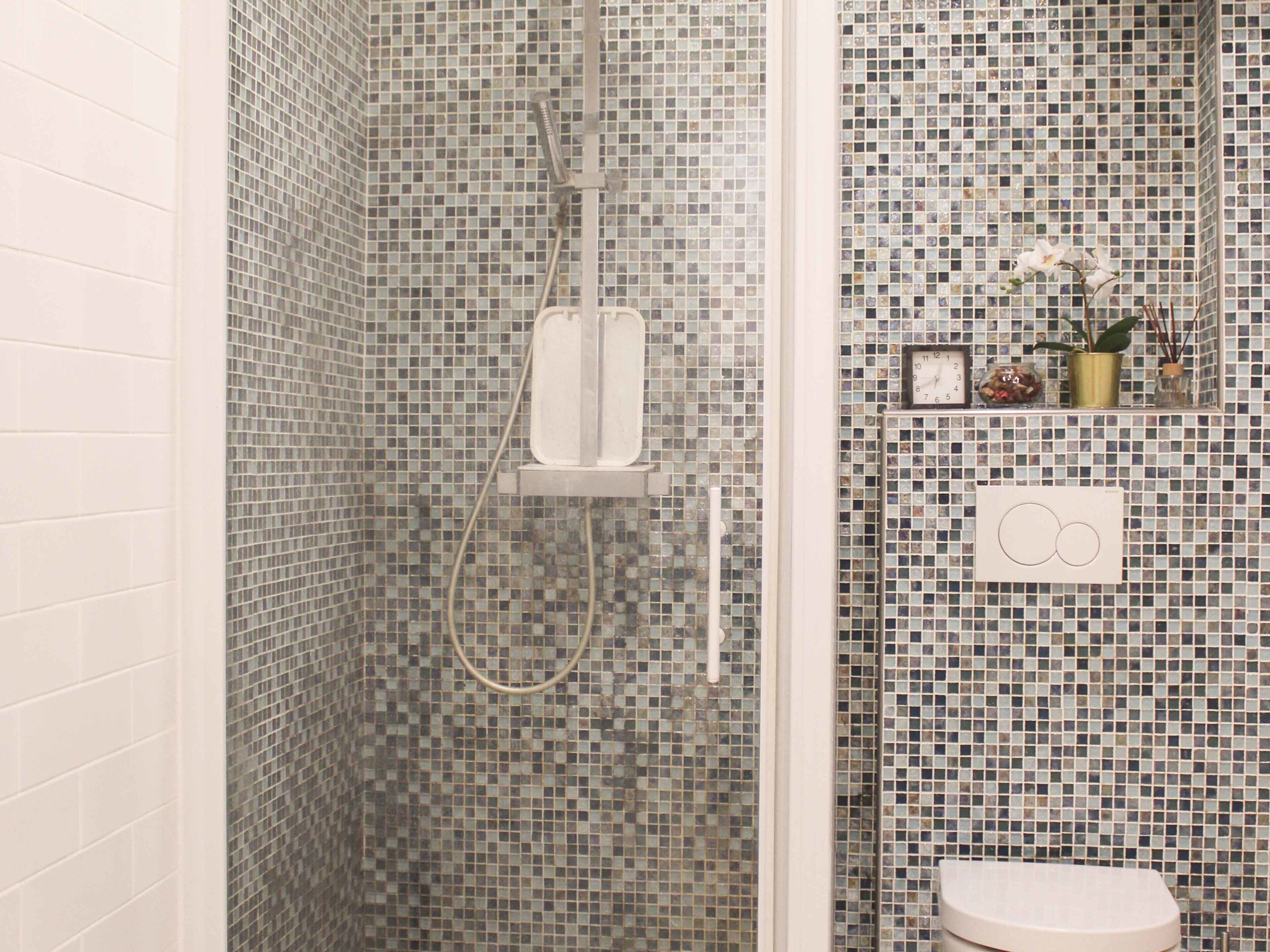 A bathroom with mosaic tile walls, a shower with a handheld showerhead, a toilet, and a shelf with decorative items.