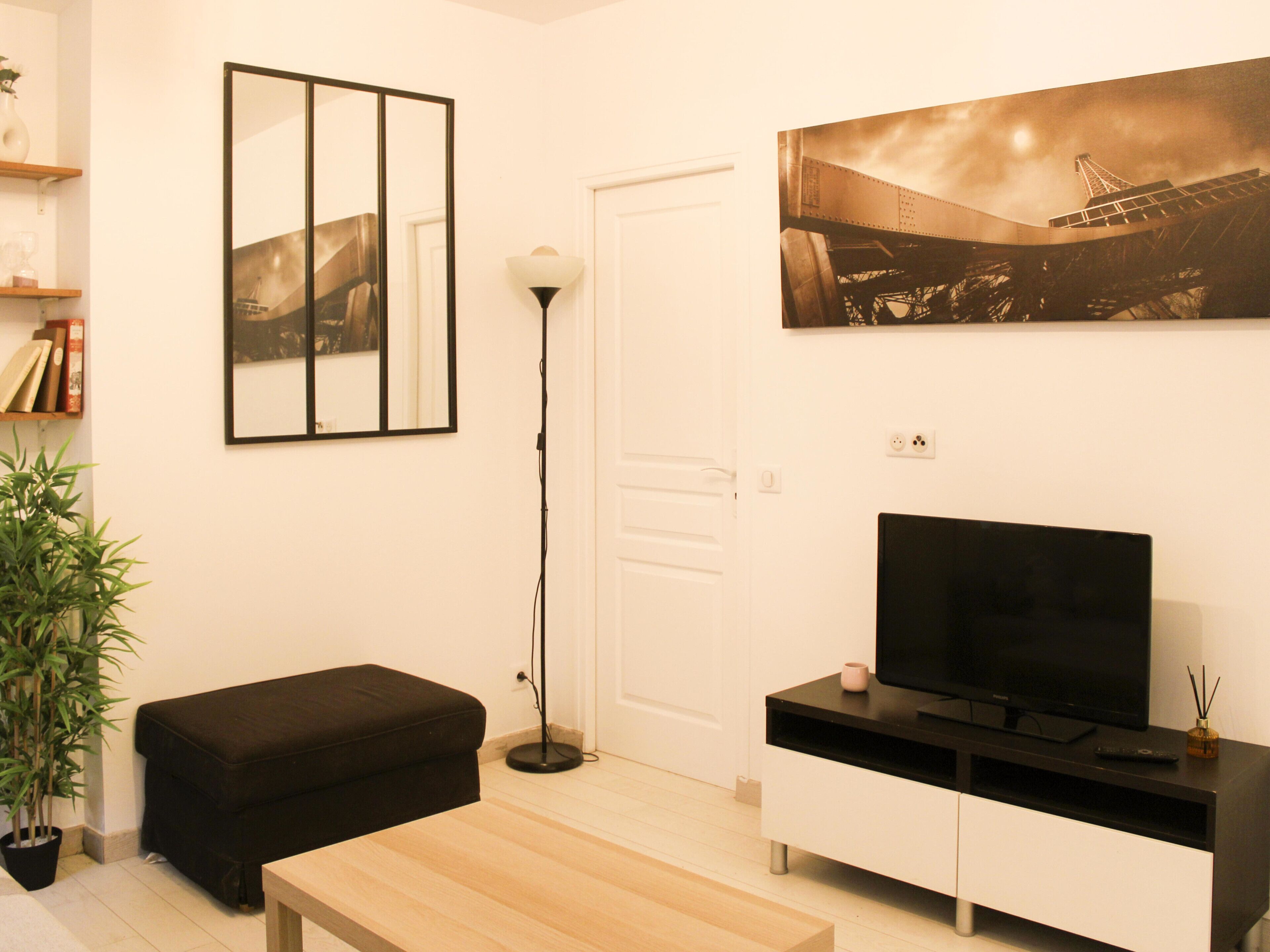 Een moderne woonkamer met een flatscreen televisie, een zwart voetenbankje, een houten salontafel, een vloerlamp en een ingelijst beeld van een brug.