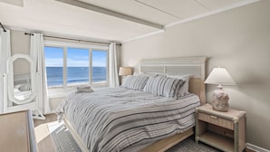 A bedroom with a large window overlooking the sea, a bed with striped bedding, a bedside table with a lamp, and a mirror. - Salty Sands 2 301 | Oceanfront w/ Balcony & Views (Ocean City)