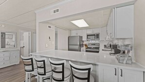 A modern kitchen with white cabinetry, a central island with bar stools, and stainless steel appliances. - Salty Sands 2 301 | Oceanfront w/ Balcony & Views (Ocean City)