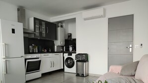 A modern kitchen with white appliances, a washing machine, and a trash bin. - Vue Exceptionelle ! Mer-port- Esplanade Oniria ! (Canet-en-Roussillon)