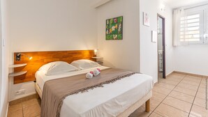 A neatly made bed with a wooden headboard, a bedside table, and a wall-mounted artwork. - Studio 102 - Le Balisier (Les Trois-Îlets)