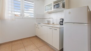 A kitchen with white cabinets, a microwave, a refrigerator, and a window with blinds. - Studio 102 - Le Balisier (Les Trois-Îlets)