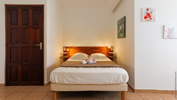 A neatly made bed with a wooden headboard, a wooden door, and two wall-mounted paintings. - Studio 102 - Le Balisier (Les Trois-Îlets)