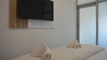 A modern hotel room with a flat-screen TV mounted on a wooden panel, a large window, and neatly made beds with rolled towels.