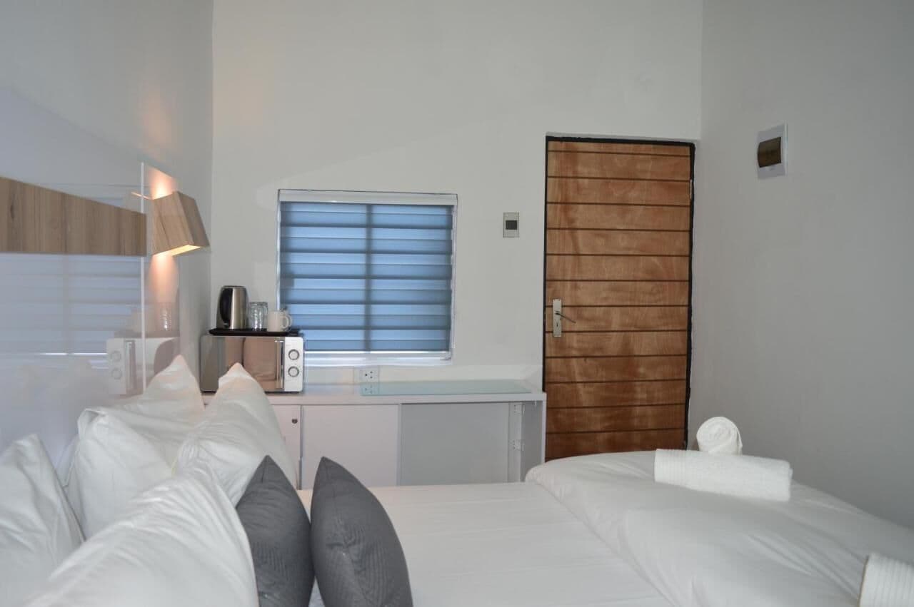 A hotel room with a bed, a desk with a kettle, a window with blinds, and a wooden door.
