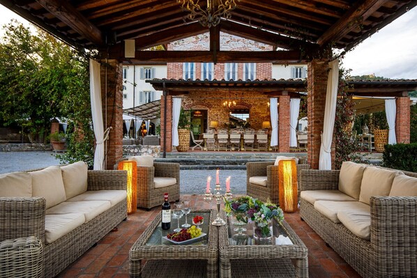 A covered outdoor seating area with wicker furniture, a table with wine glasses and a bottle, and a stone building in the background.