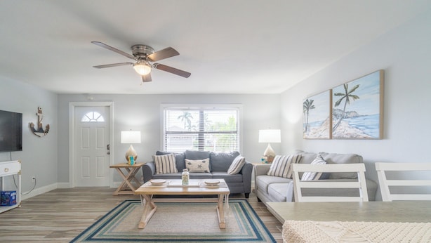 A living room with a ceiling fan, a TV, a sofa, a coffee table, and a painting of a beach scene.