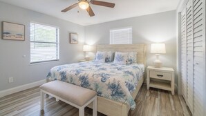 A bedroom with a bed, a bench, two bedside tables with lamps, a ceiling fan, and two windows with blinds.