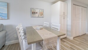 A dining area with a wooden table, white chairs, a macrame runner, and a coastal painting on the wall.