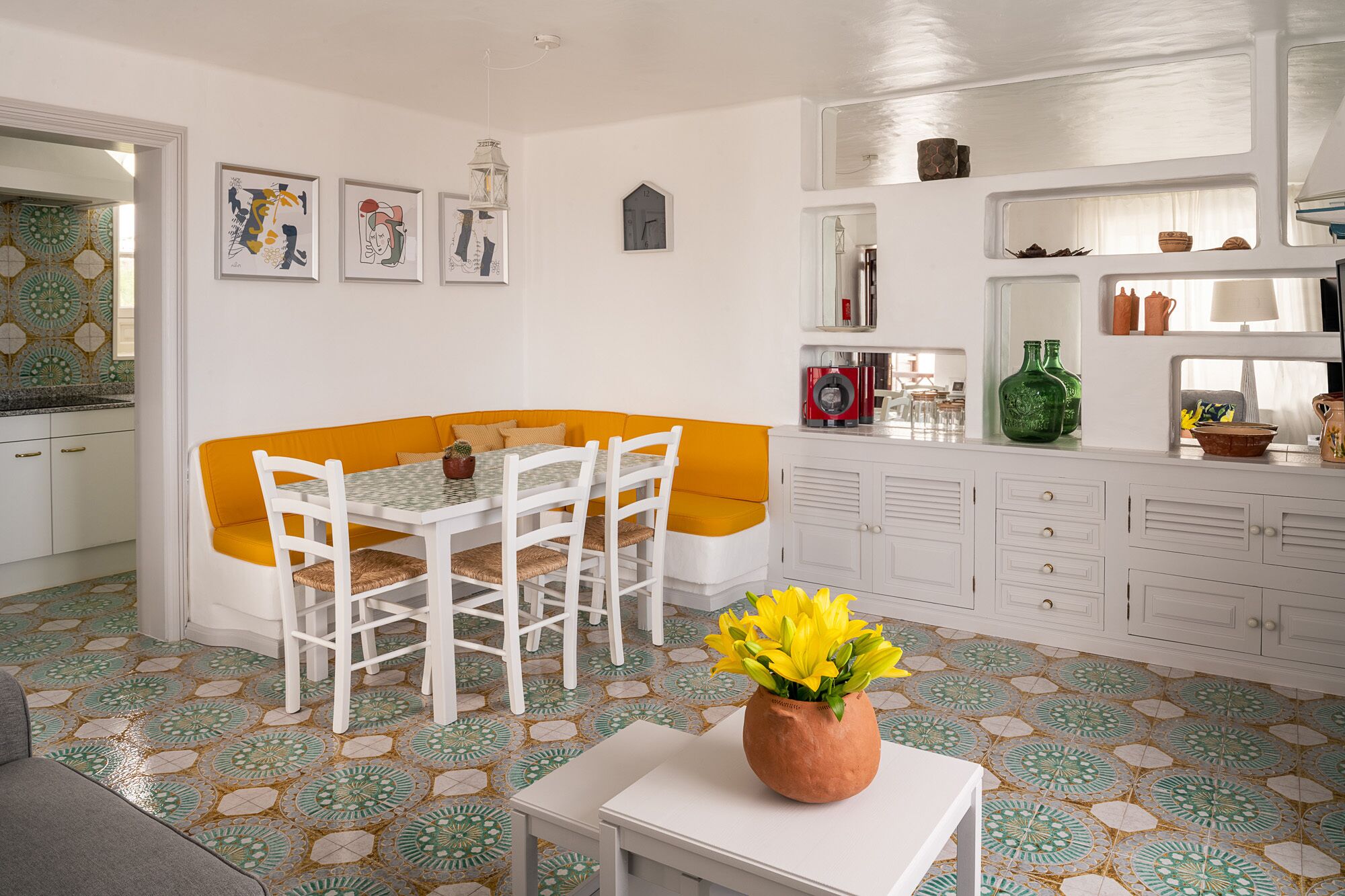 A dining area with a white table and chairs, a corner sofa with orange cushions, and a vase of yellow flowers on a small table.