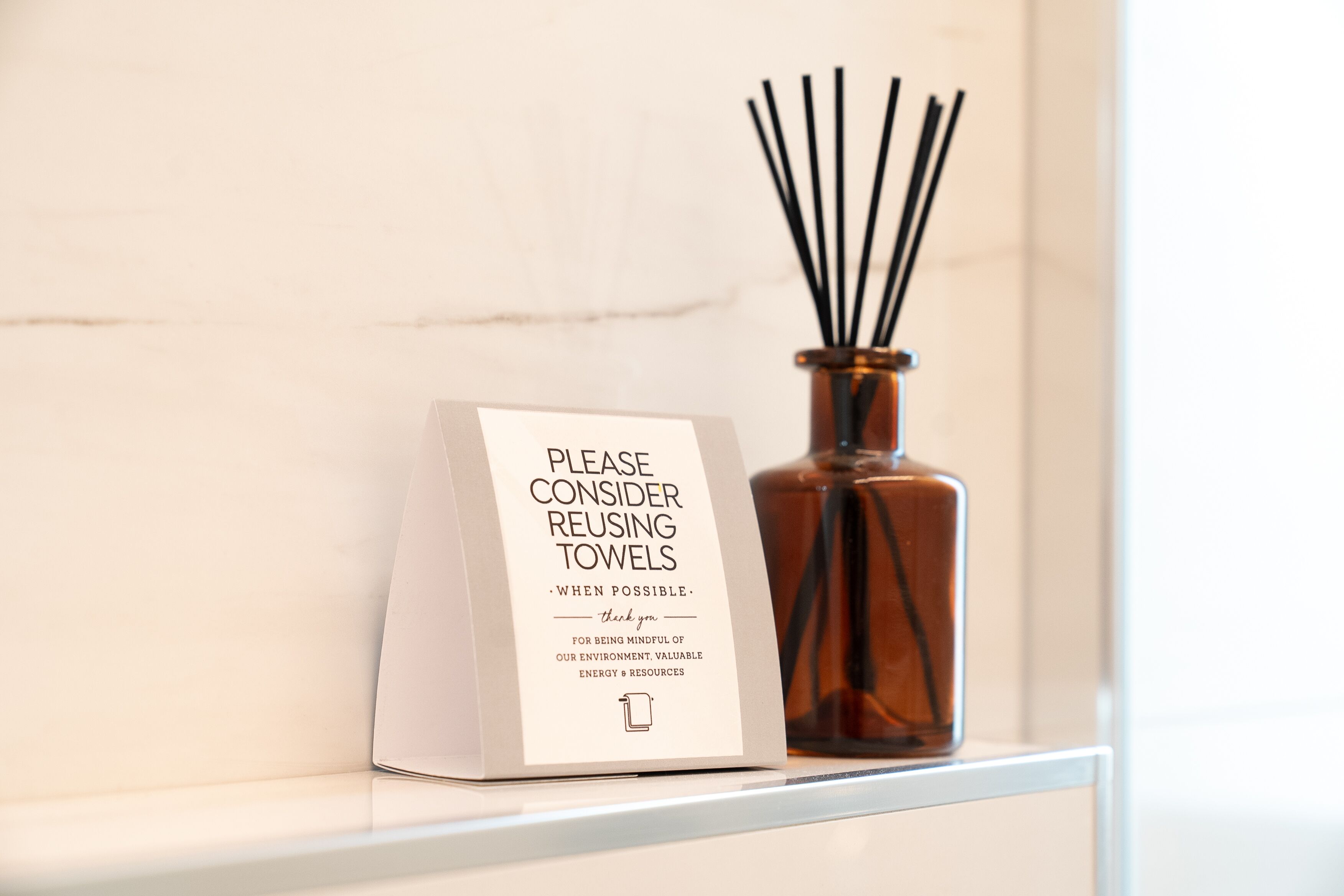 Une pancarte suggérant de réutiliser les serviettes pour des raisons environnementales est placée à côté d’un vase marron contenant des tiges de roseau.