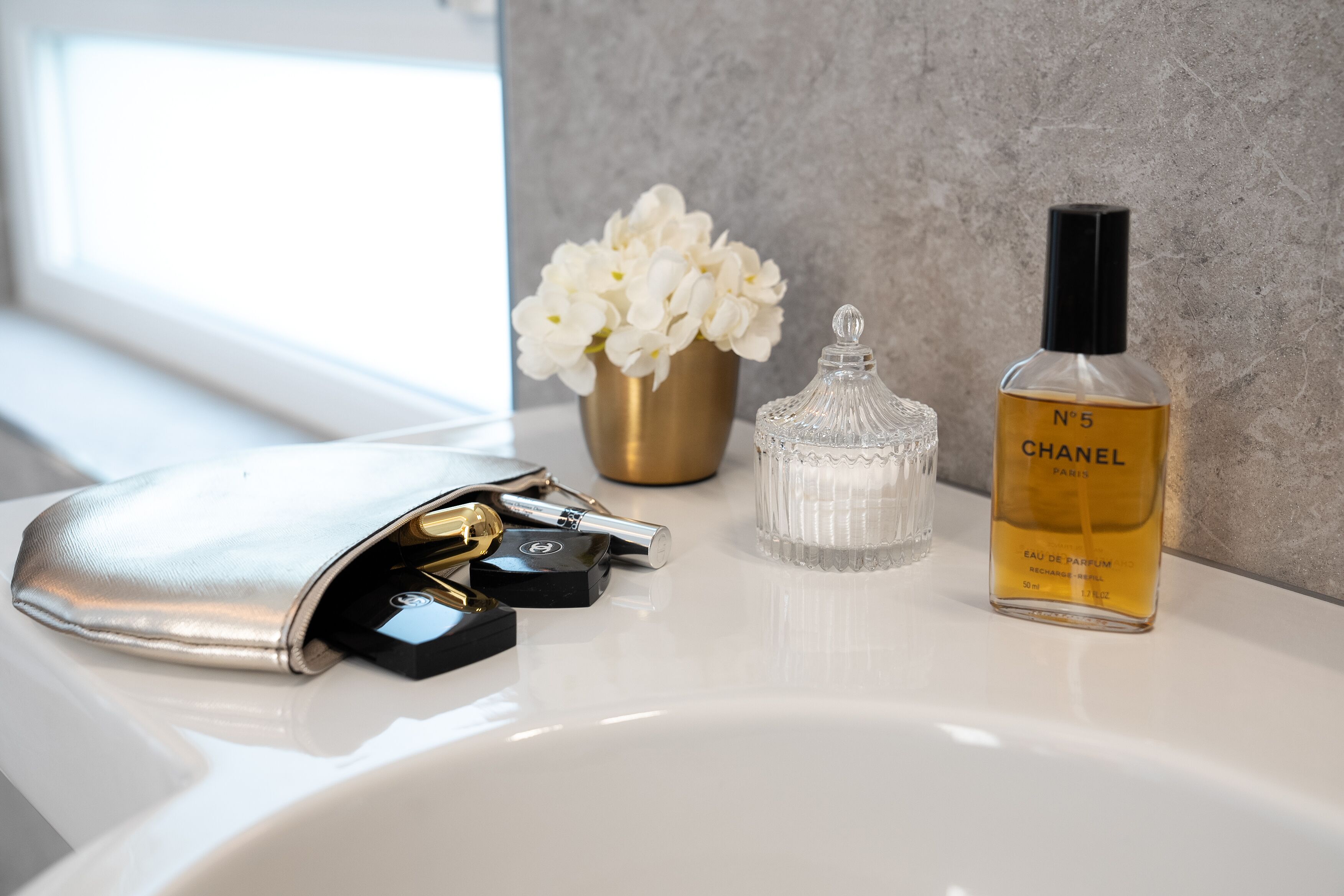 A bathroom vanity with a Chanel perfume bottle, a gold vase with white flowers, and a silver toiletry bag.
