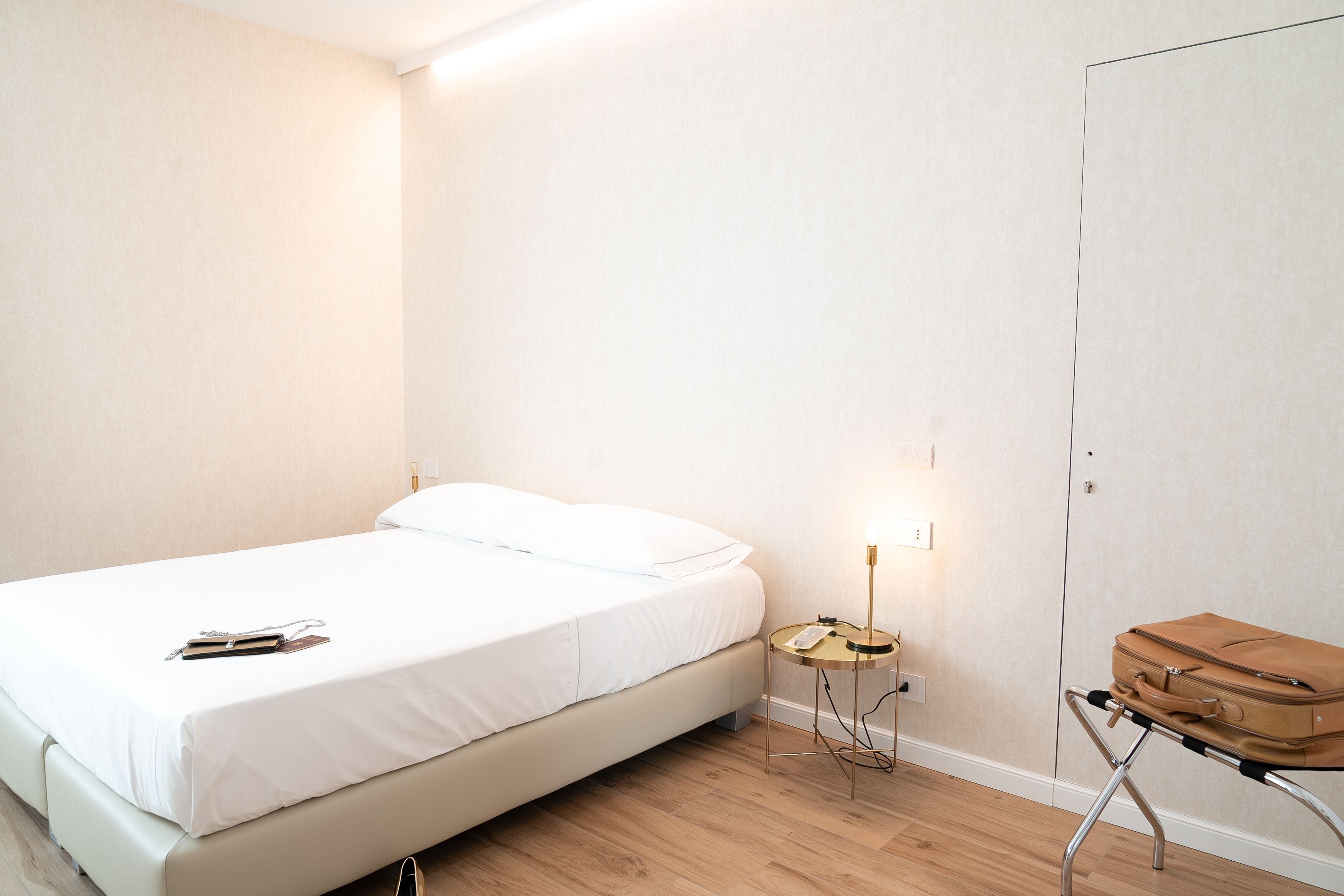 A neatly made bed with a white sheet and a book beside it. There is a small bedside table with a lamp and a suitcase on a stand.