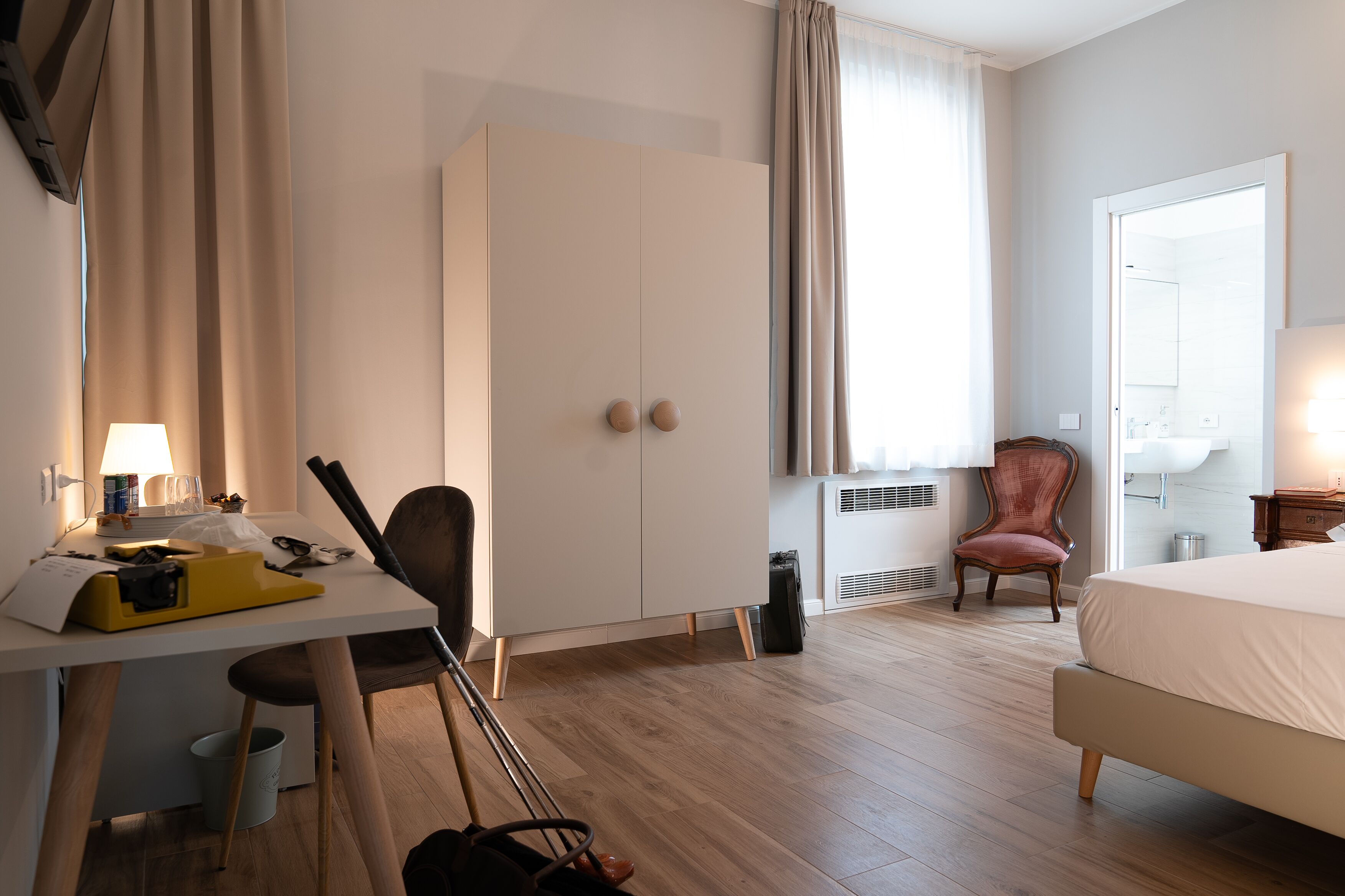 Quarto de hotel com cama, escrivaninha com luminária, guarda-roupa, cadeira e banheiro com espelho e pia.