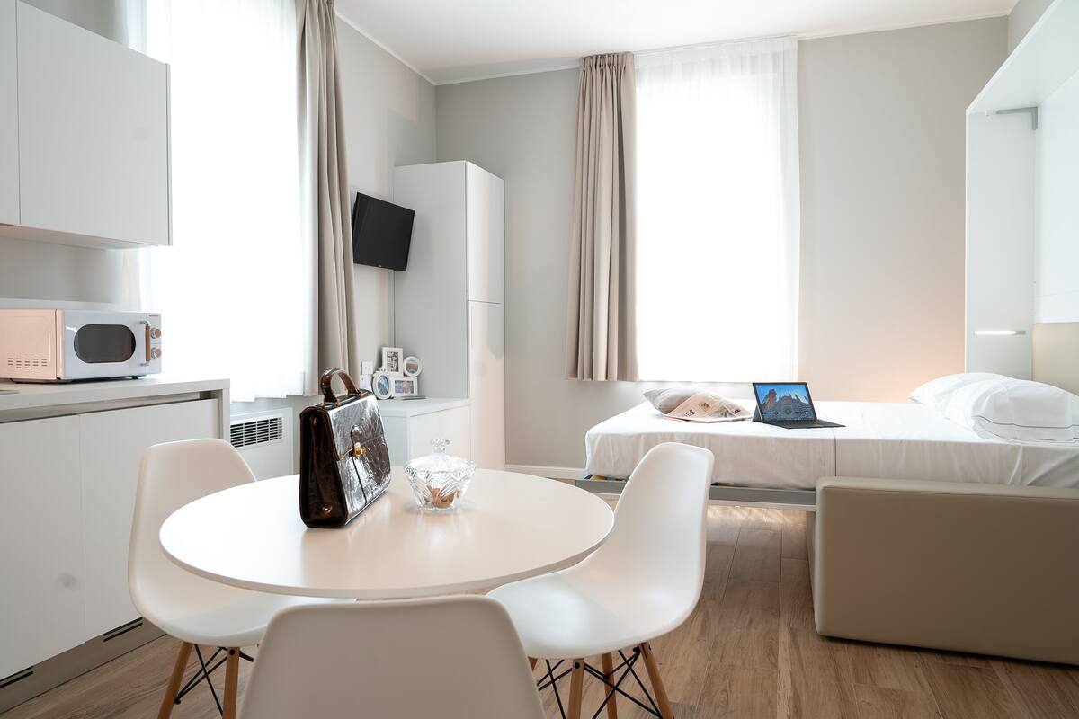 A modern hotel room with a dining area, a bed with a laptop, and a TV mounted on the wall.