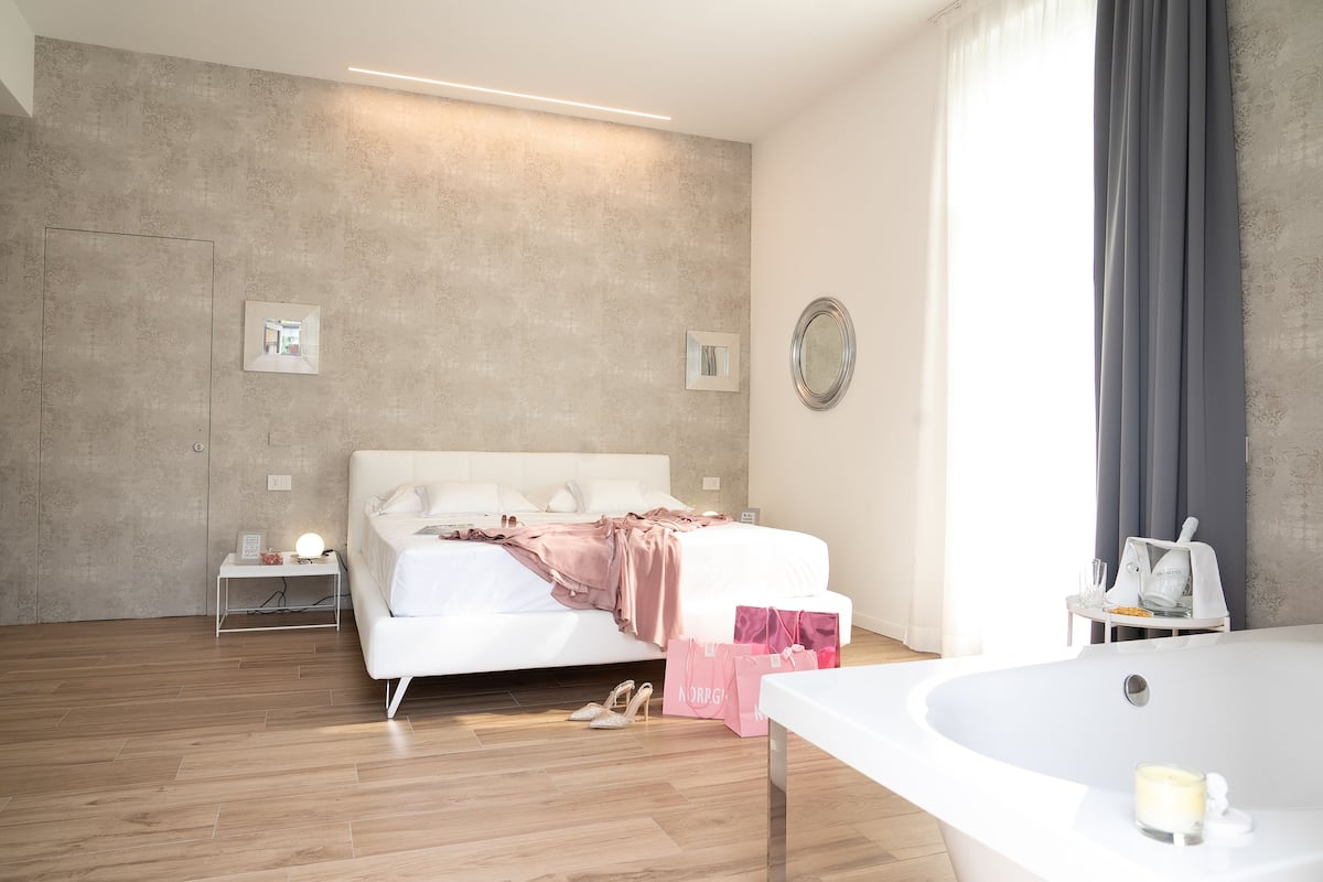 A modern bedroom with a large bed, a bedside table with a lamp, a small table with a mirror, and a bathtub with a towel and candle.