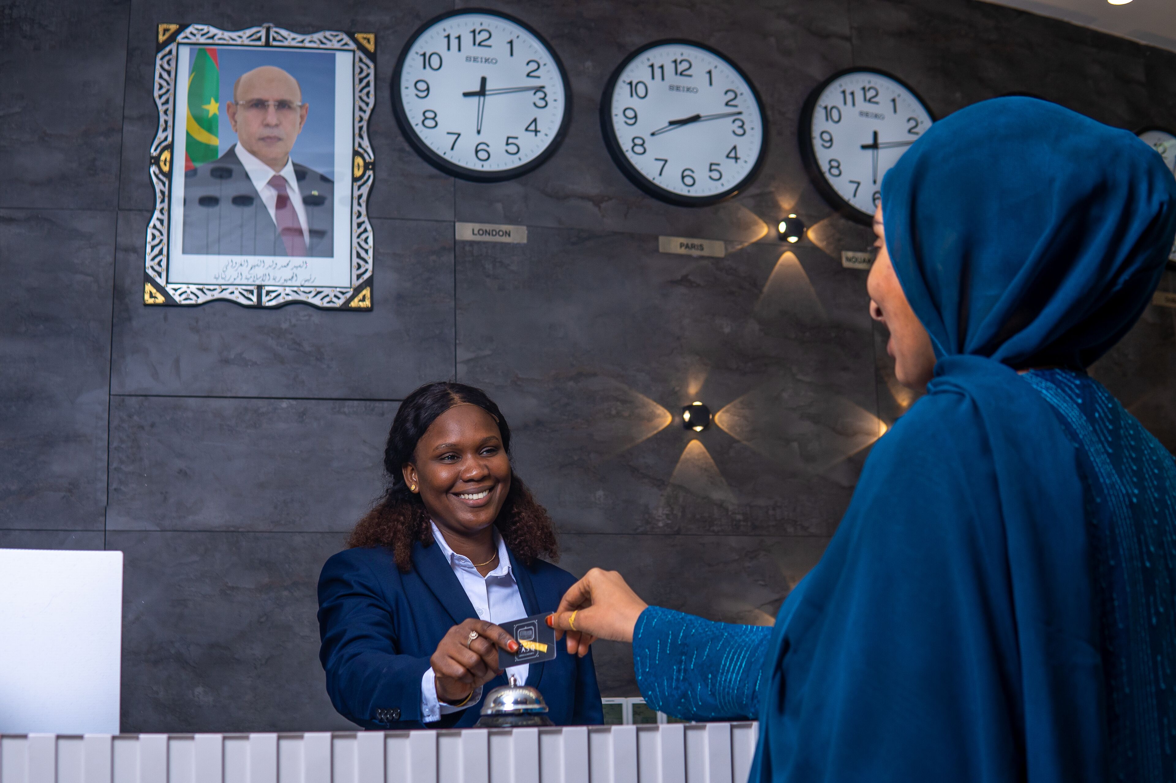 A reception desk with clocks labeled 'LONDON' and 'PARIS', a framed portrait, and a person handing over a card.
