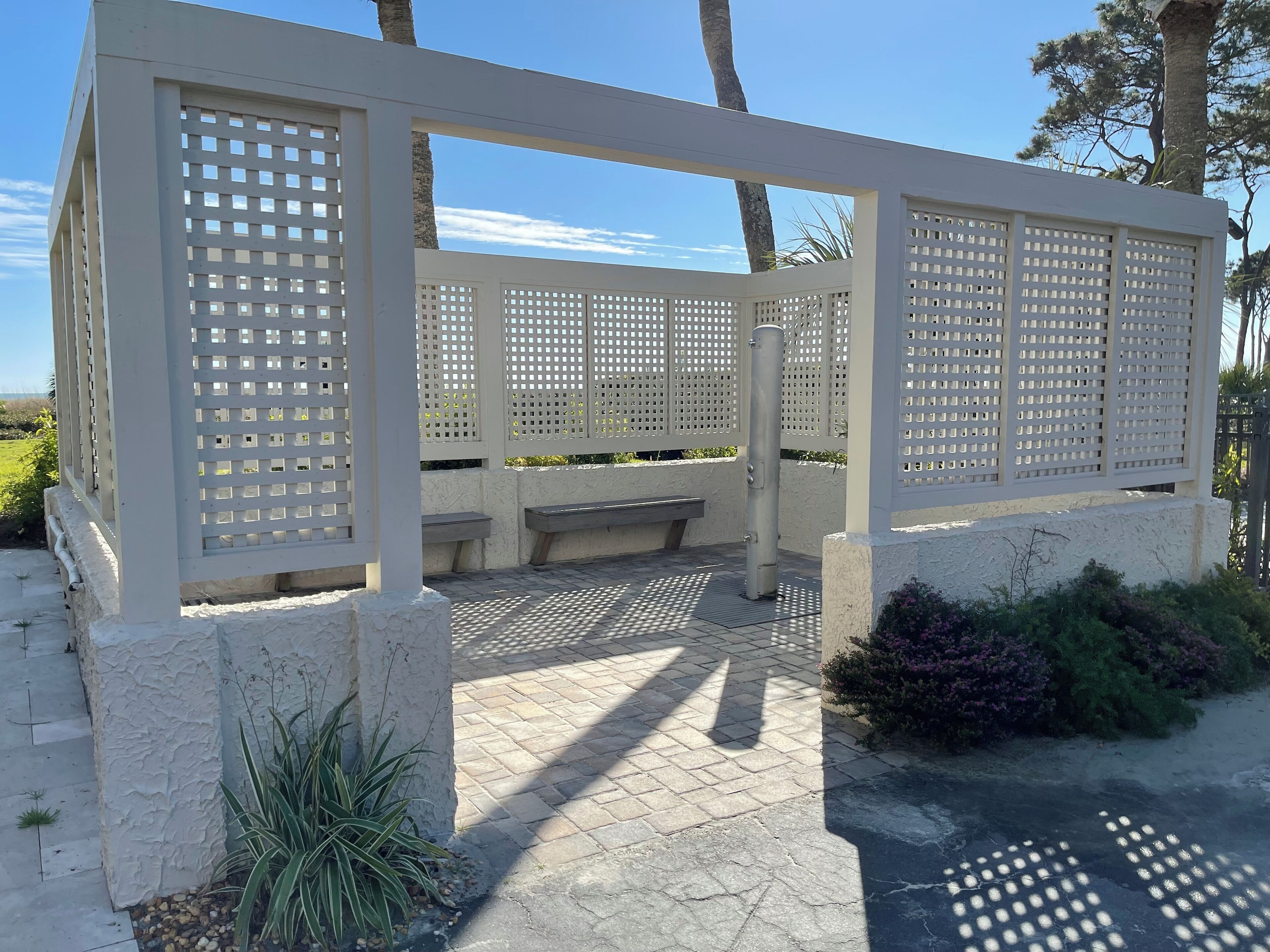 A covered outdoor seating area with a white lattice structure, stone flooring, and a bench.
