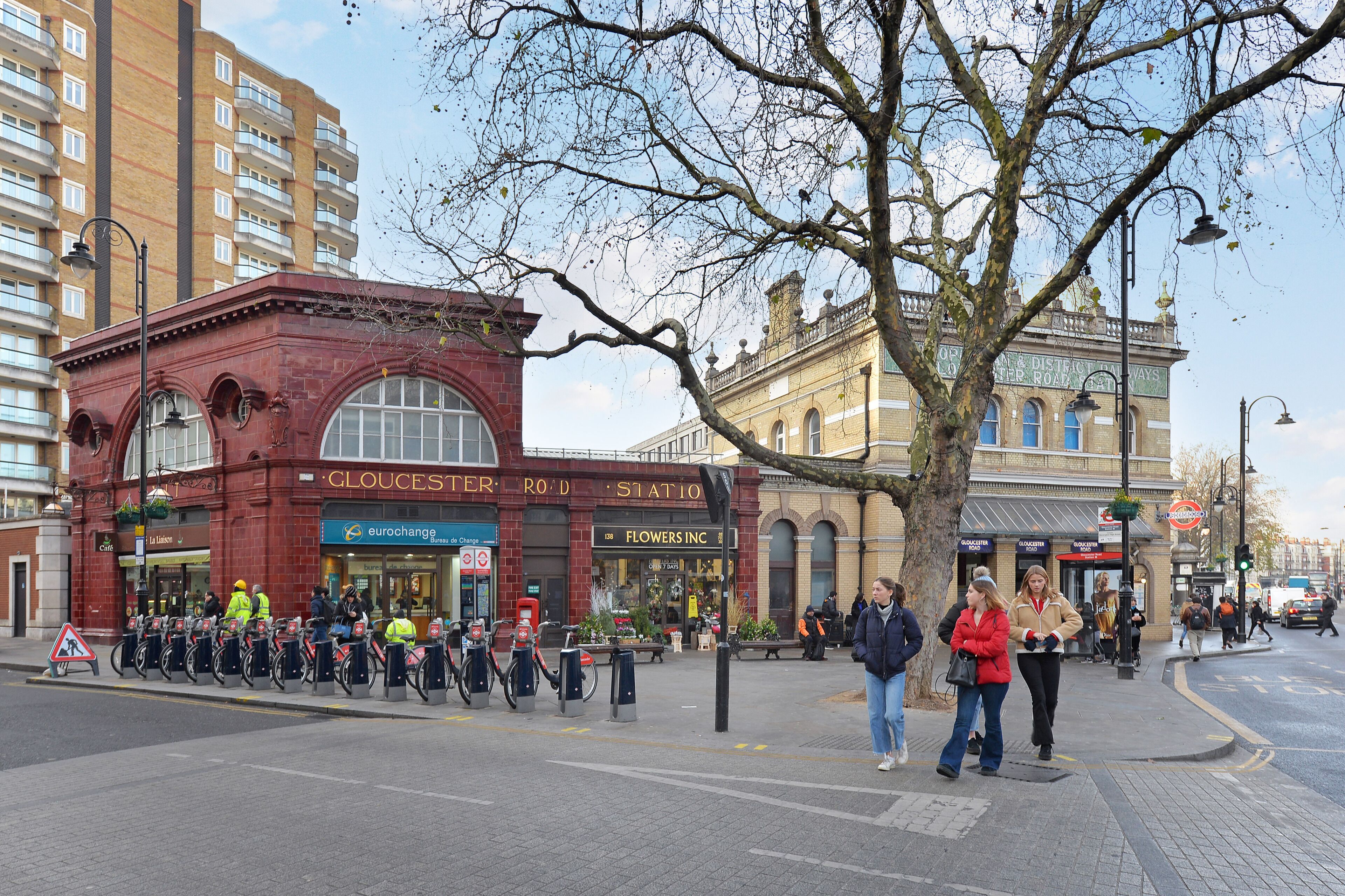 Une scène de rue avec un bâtiment rouge portant l’inscription « GLOUCESTER ROAD STATION » et un arbre au premier plan.