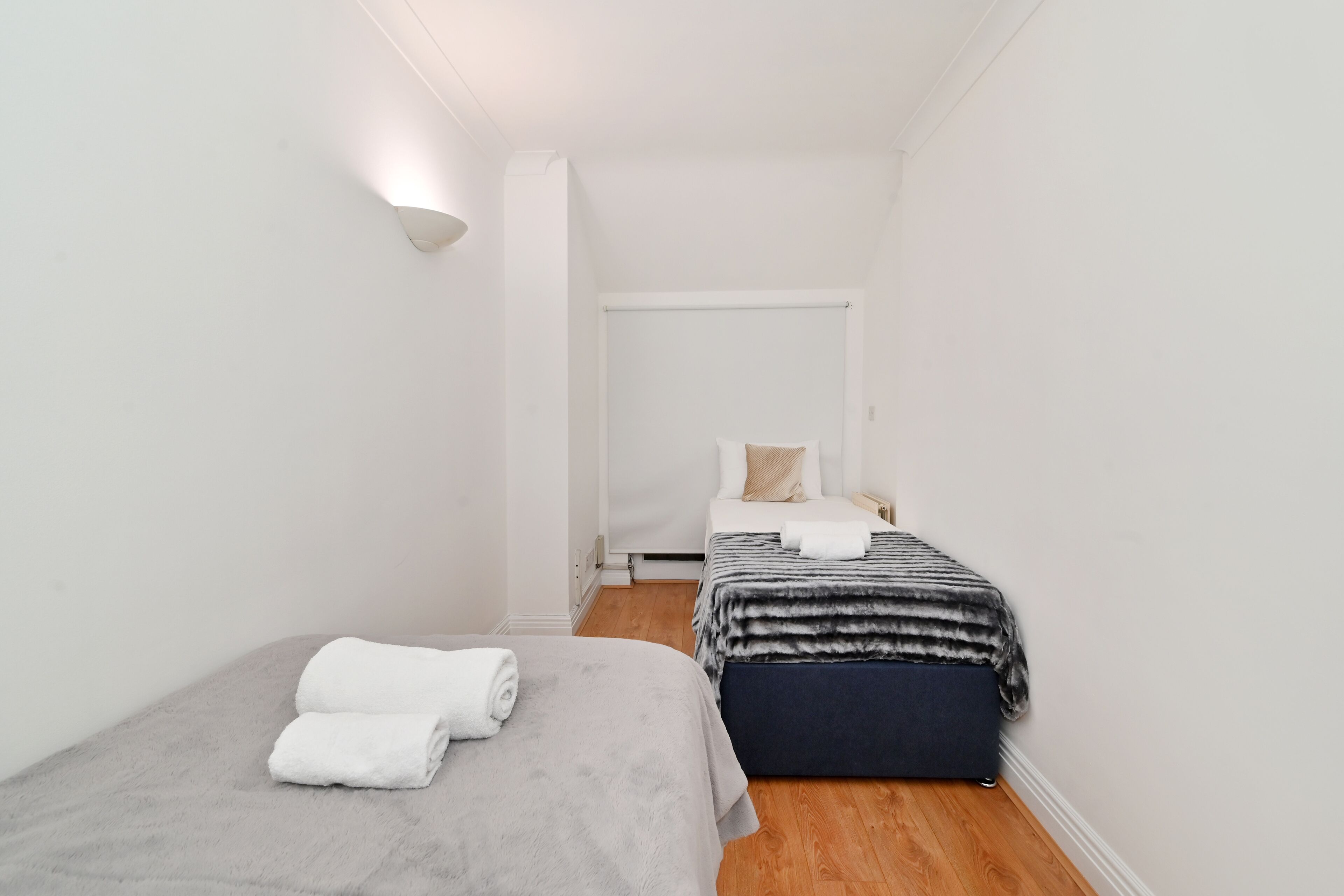Una habitación pequeña y limpia con dos camas, una con una manta gris y la otra con una manta a rayas.