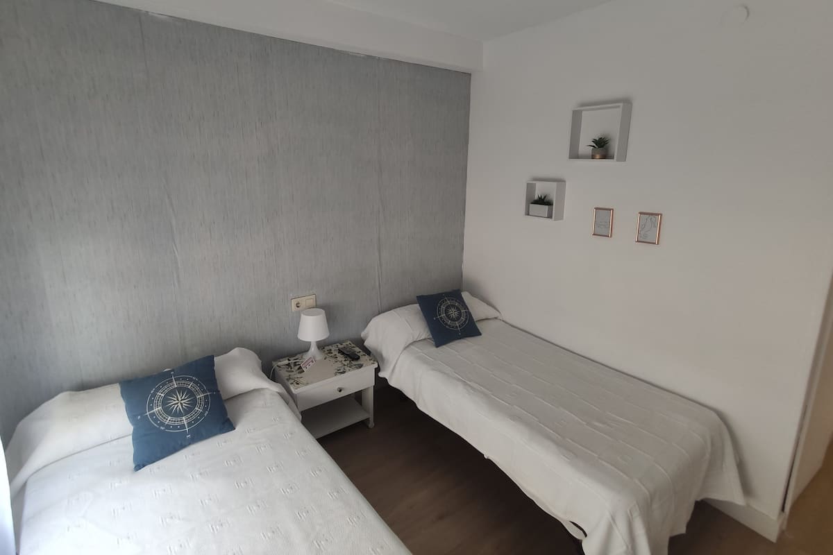 A bedroom with two single beds, a nightstand, a lamp, and a small shelf with a plant.