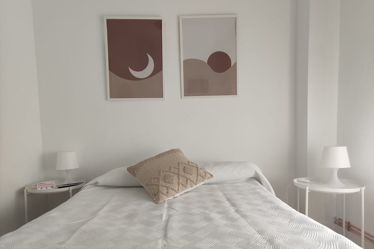A neatly made bed with a patterned white bedspread, two wall-mounted artworks depicting a crescent moon and a sun, and two bedside tables with lamps.