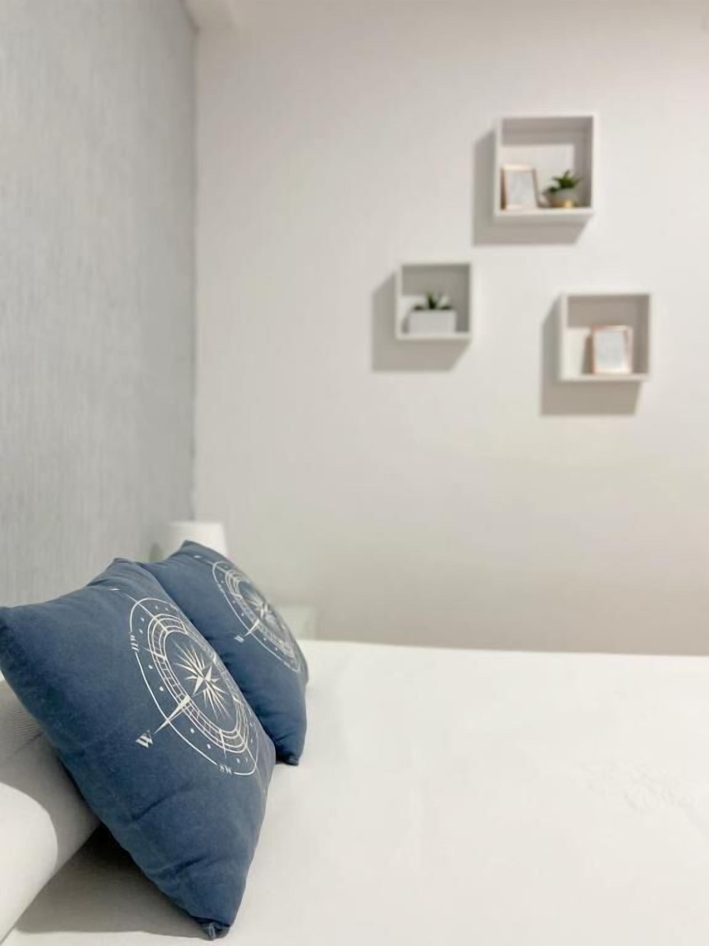 Quarto com uma cama branca, dois travesseiros azuis com desenho branco e três mesas de cabeceira brancas com objetos pequenos.
