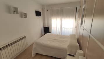 A modern hotel room with a large bed, a flat-screen TV mounted on the wall, a radiator, and a window with sheer curtains.