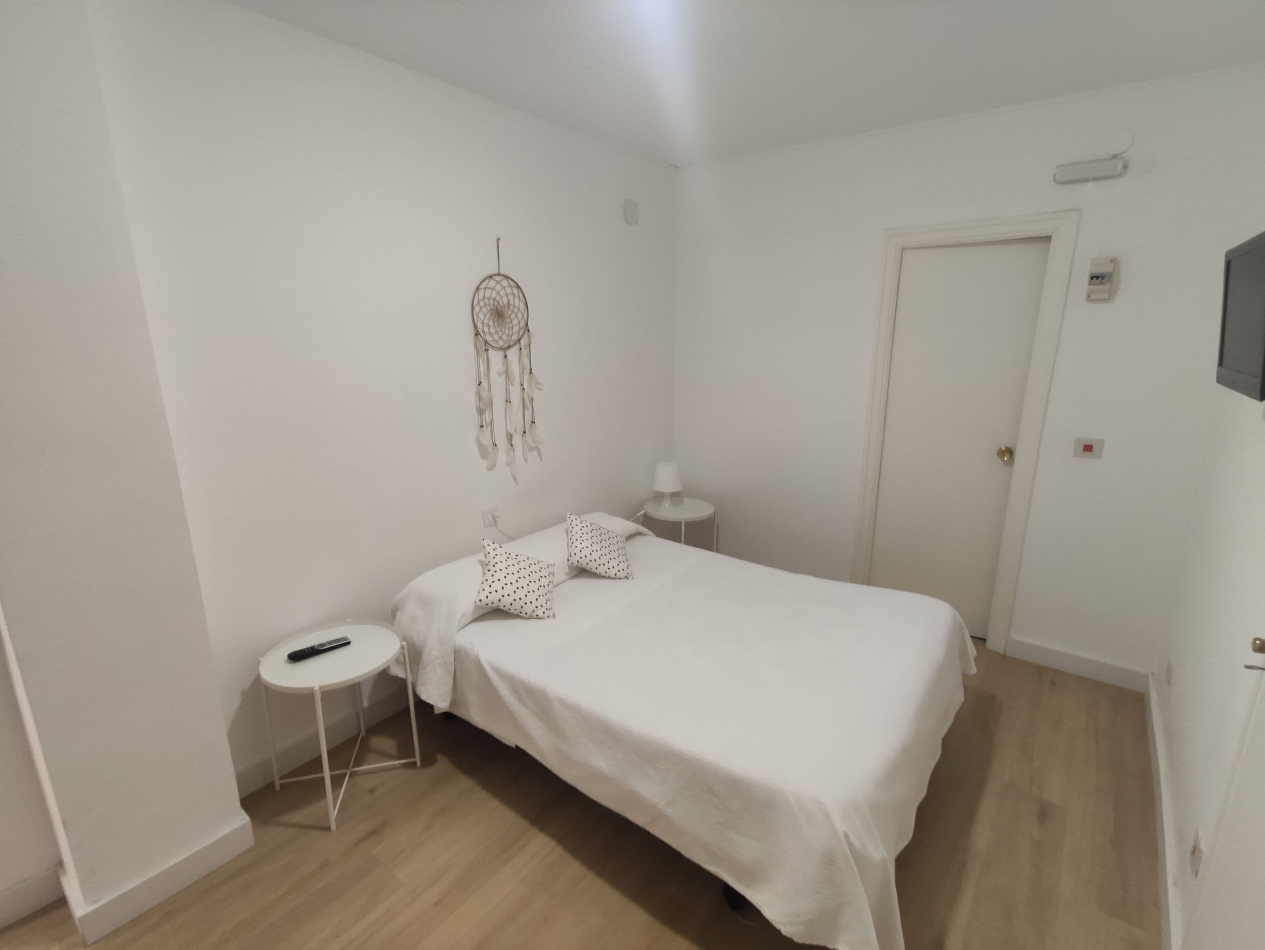 Une chambre à coucher minimaliste, avec un lit simple, une table de chevet, une petite table ronde et un attrape-rêves accroché au mur.