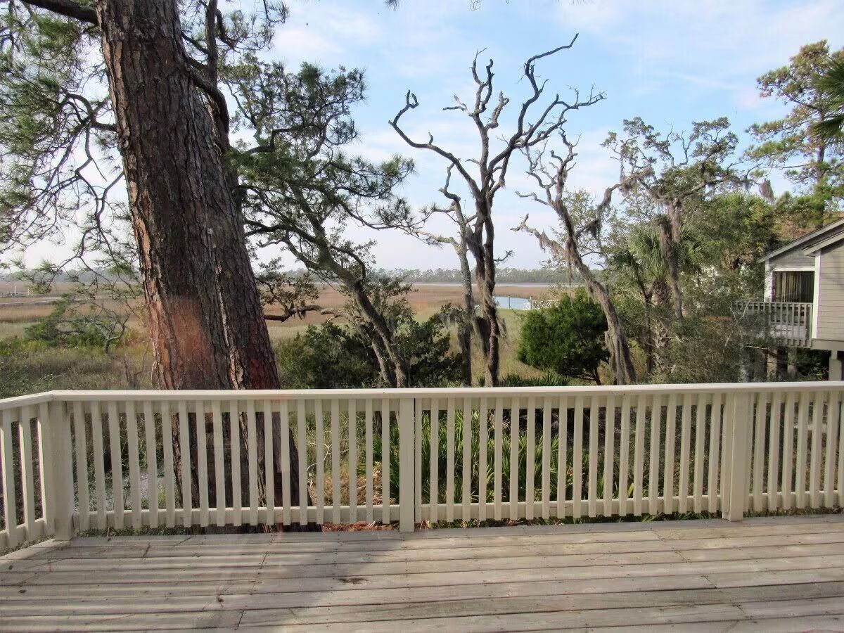 Een houten terras met een witte balustrade, omringd door bomen en een huis op de achtergrond.