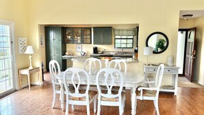 Een eetgedeelte met een witte tafel en stoelen, een keuken met groene kasten, en een raam met gordijnen.