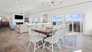 A spacious dining area with a large table and white chairs, a chandelier, and a view of the ocean through large sliding doors. - Luxury Oceanfront Retreat | 6 Ensuite Bedrooms | Sleeps 14 | Golf Cart Included | Steps to Beach Club (Fripp Island)