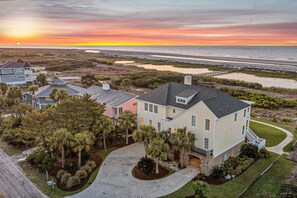 A large, multi-story house with a spacious driveway and a well-maintained lawn, located near a beach with a sunset view.