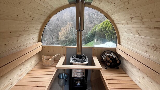 A wooden cabin with a central wood stove, benches, and a window with a view of trees. - Modern Villa with heated jacuzzi,sauna,panoramic view,close to Tuscany (Pergoloso)
