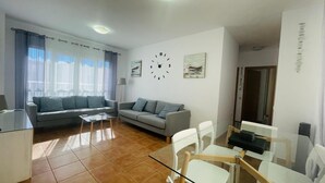 Un salon moderne avec deux canapés gris, une table basse en verre et un coin repas comprenant une table et des chaises blanches.