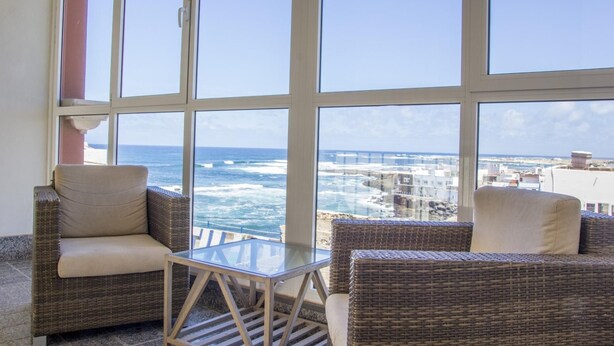 Un balcon doté de meubles en osier et d’une table en verre, offrant une vue sur la côte, avec ses bâtiments et la mer.