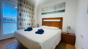 Une chambre d’hôtel avec un grand lit, une tête de lit en bois, une table de chevet avec une lampe et une vue sur la mer par la fenêtre.