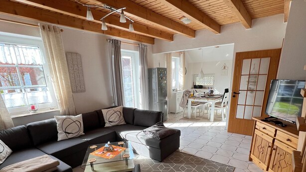 Un salon moderne avec un canapé, une table basse et un téléviseur. On aperçoit une cuisine au fond et un coin repas.