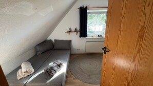 Une pièce chaleureuse avec un canapé gris, une porte en bois, une fenêtre avec des rideaux et une petite étagère ornée d’objets décoratifs.